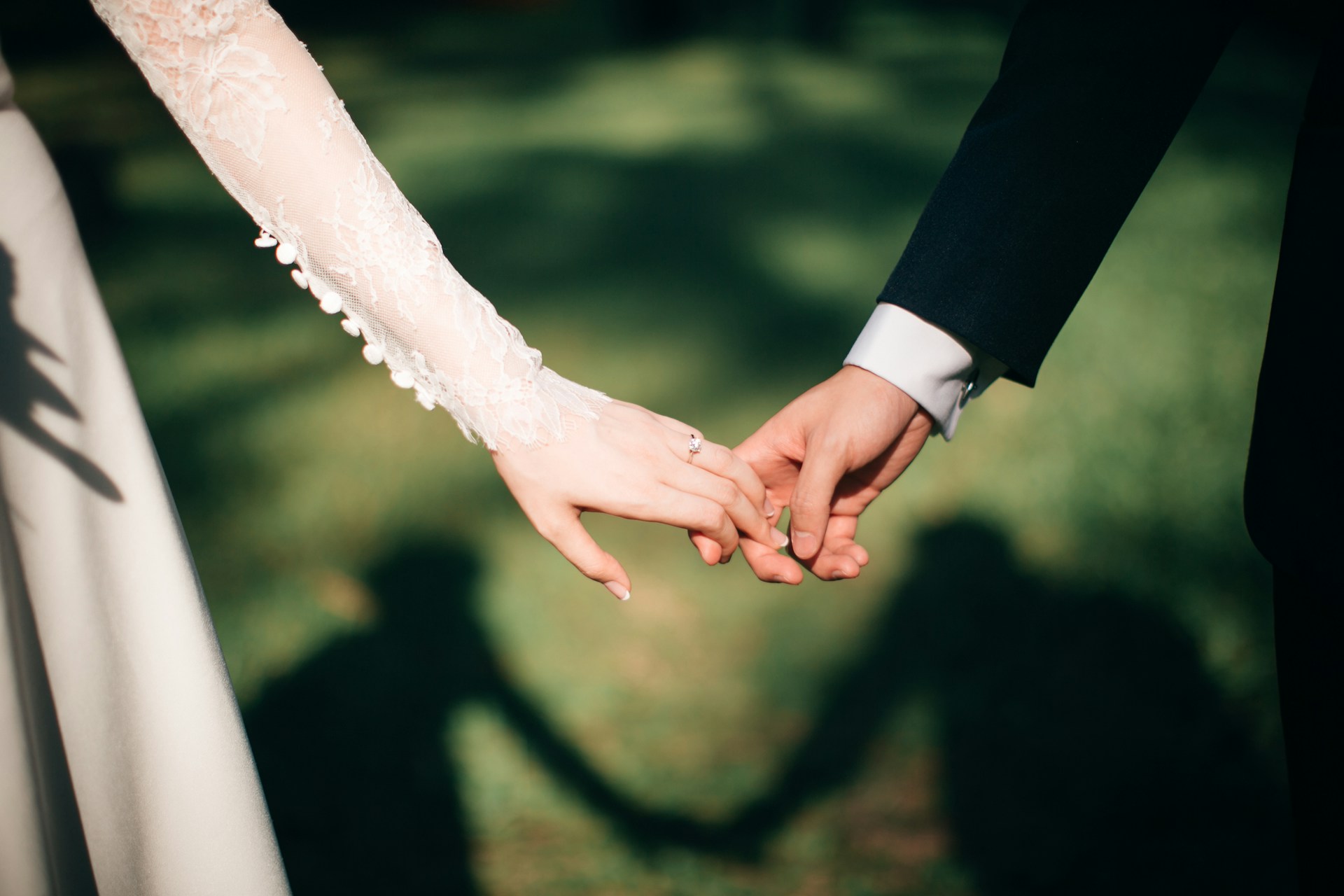 Close-up of a bride wearing a lace sleeve and a groom holding hands outdoors.